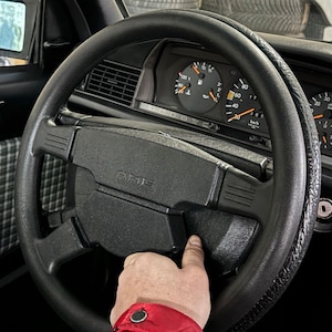 以下が含まれることがあります： 中央に「AMG」の文字が入った黒いステアリングホイール。ステアリングホイールの後ろにはダッシュボードのゲージが見えます。手がステアリングホイールに触れています。車の内装は黒です。