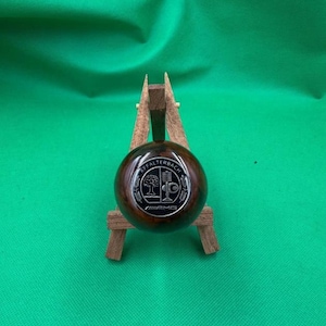 May include: A dark brown, spherical object with a silver emblem featuring a tree and the text "AFFALTERBACH" and "AMG". The object is displayed on a small wooden stand against a green background.