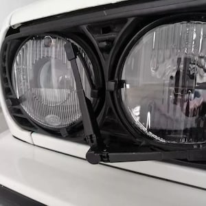 May include: Close-up of a white car's front end, showcasing two round headlights with a black wiper system. The headlights are clear with a ribbed pattern, and the car's bumper is white.