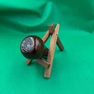 Puede incluir: Un objeto esférico, brillante y marrón oscuro, con un emblema plateado, apoyado en un pequeño soporte de madera. El soporte está hecho de finas piezas de madera de color marrón claro. El fondo es verde brillante liso.