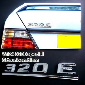 May include: A silver 320E boot emblem for a Mercedes-Benz W124. The emblem is made of metal and has a polished finish. The emblem is attached to a black surface.