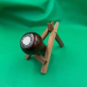 May include: A dark brown, cylindrical object with the text "AFFALTERBACH" and "AMG" in a silver circle, resting on a small wooden stand. The stand is made of light brown wood and has a simple, geometric design. The background is a solid green.