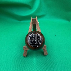 May include: A dark brown, round object with a silver emblem featuring the text "Affalterbach" and "AMG". The object is displayed on a small wooden stand against a green background. The emblem has a tree and a logo.