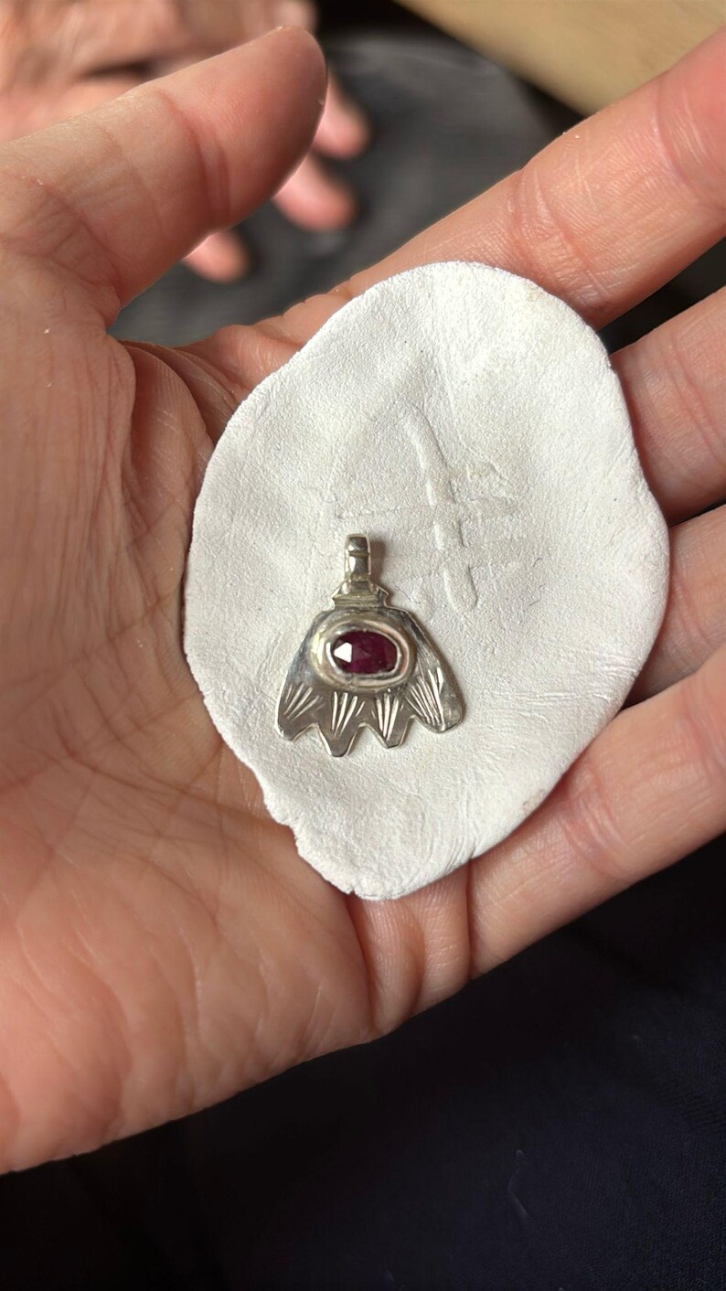 May include: A silver pendant featuring a red gemstone in the centre. The pendant is shaped like a stylised flower with five petals.