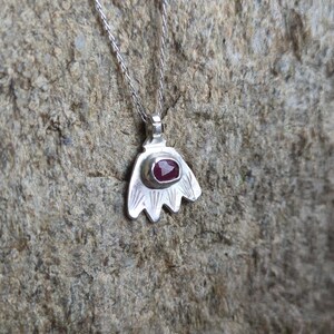May include: A silver necklace with a pendant shaped like a stylised flower. The pendant has a red gemstone in the centre.