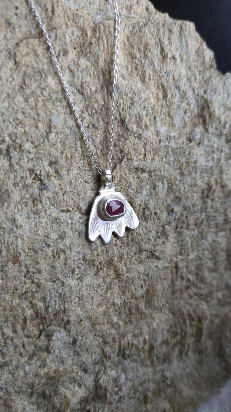 May include: A silver necklace with a pendant shaped like a stylised flower. The pendant has a red gemstone in the centre.