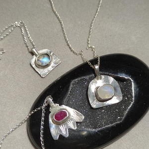 May include: Three silver necklaces with different pendants. The pendants feature a square with a clear stone, a rounded shape with a white stone, and a flower-shaped pendant with a red stone.