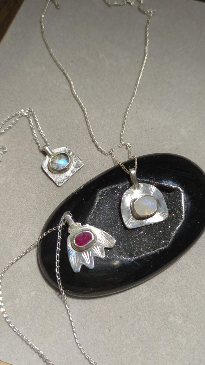 May include: Three silver necklaces with different pendants. The pendants feature a square with a clear stone, a rounded shape with a white stone, and a flower-shaped pendant with a red stone.