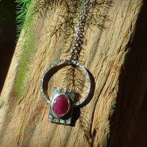 May include: A silver pendant necklace with a large, oval, deep red gemstone set in a textured silver frame. The pendant hangs from a delicate silver chain. The necklace is displayed on a rustic wooden surface, with green foliage in the background.