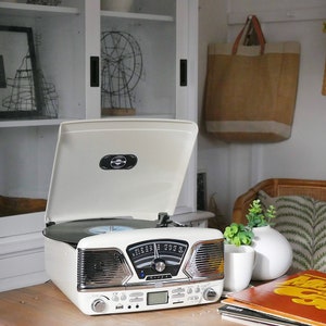 May include: A cream-coloured vintage-style record player with an open lid, revealing a black vinyl record. The player has a silver-toned front panel with a radio dial and speaker grilles. A small white vase with greenery and a stack of vinyl records are nearby.