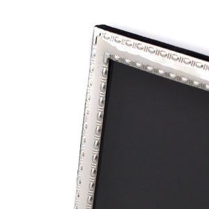 May include: A rectangular silver picture frame with a black interior. The frame has a decorative beaded border and a black backing. The frame is angled slightly, showing its side profile. The frame is made in England.