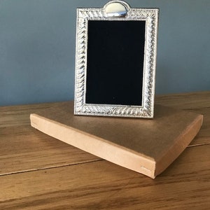 May include: A silver-colored picture frame with a black interior, resting on a brown cardboard box. The frame has a decorative border and a rounded top. The box is on a wooden surface.
