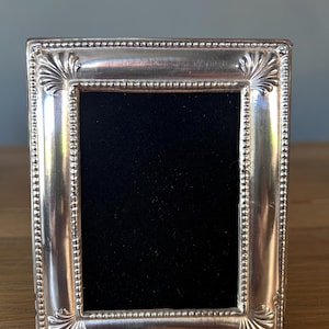 May include: A rectangular silver picture frame with a polished metallic finish, featuring a beaded inner border and decorative shell-like motifs at each corner. The center of the frame contains a solid black rectangular insert.