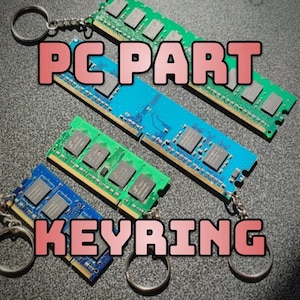 May include: Four different coloured RAM sticks with keyrings attached. The RAM sticks are green, blue, and two shades of blue. The text "PC PART KEYRING" is written in pink letters on a grey background.