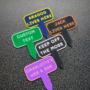 May include: Five colourful garden markers with different text. The markers are shaped like speech bubbles and have black, blue, green, orange, and pink backgrounds. The text on the markers reads: "Aragog Lives Here", "Custom Text", "Jack Lives Here", "Keep Off The Moss", and "Charlotte's Web & Bar".