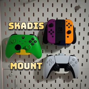 May include: Three different video game controllers mounted on a white pegboard wall. The controllers are green, purple and orange, and white. The text "SKADIS MOUNT" is written in yellow letters on the wall.