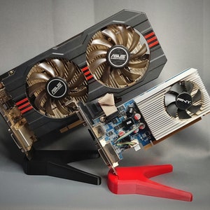 May include: Two black graphics cards with fans and red accents, and a white graphics card with a fan, are displayed on a black stand and a red stand.