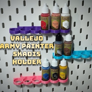 May include: A set of acrylic paint bottles organised on a wall-mounted shelf. The shelf is made of 3D printed plastic and has a purple, blue, and pink colour scheme. The bottles are labelled "Vallejo Army Painter Warpaints" and feature various colours, including red, pink, white, grey, brown, blue, black, and yellow. The text "Vallejo Army Painter Skadis Holder" is visible in the image.