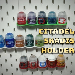 May include: A set of Citadel paint pots organised on a white pegboard wall using a 3D printed SKADIS holder. The holder is green and red. The paint pots are labelled with the Citadel Colour brand name.