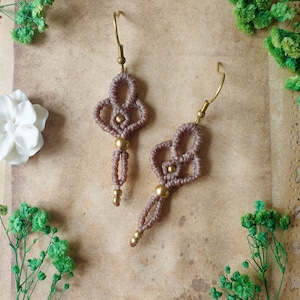 Puede incluir: Un par de pendientes de macramé marrón con cuentas doradas y ganchos. Los pendientes cuelgan de un trozo de papel marrón con flores blancas y ramitas verdes.
