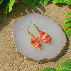 May include: A pair of handmade earrings with a leaf shape, featuring a colorful pattern of red, orange, and yellow thread. The earrings are made with gold-colored metal hooks.