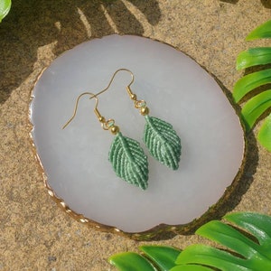 May include: A pair of green leaf-shaped earrings with gold hooks. The earrings are made of macrame and have a delicate, bohemian style.
