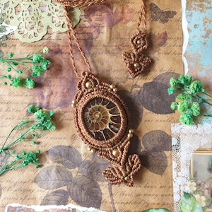 May include: A brown cord necklace with a gold and brown macrame pendant featuring a fossilized ammonite. The necklace is displayed on a background of vintage paper with floral designs.
