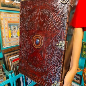 May include: Large, dark brown leather-bound book with a prominent embossed pentagram design. The cover features intricate Celtic knotwork and a central blue gemstone. Metal clasps and corner protectors add to the antique aesthetic.