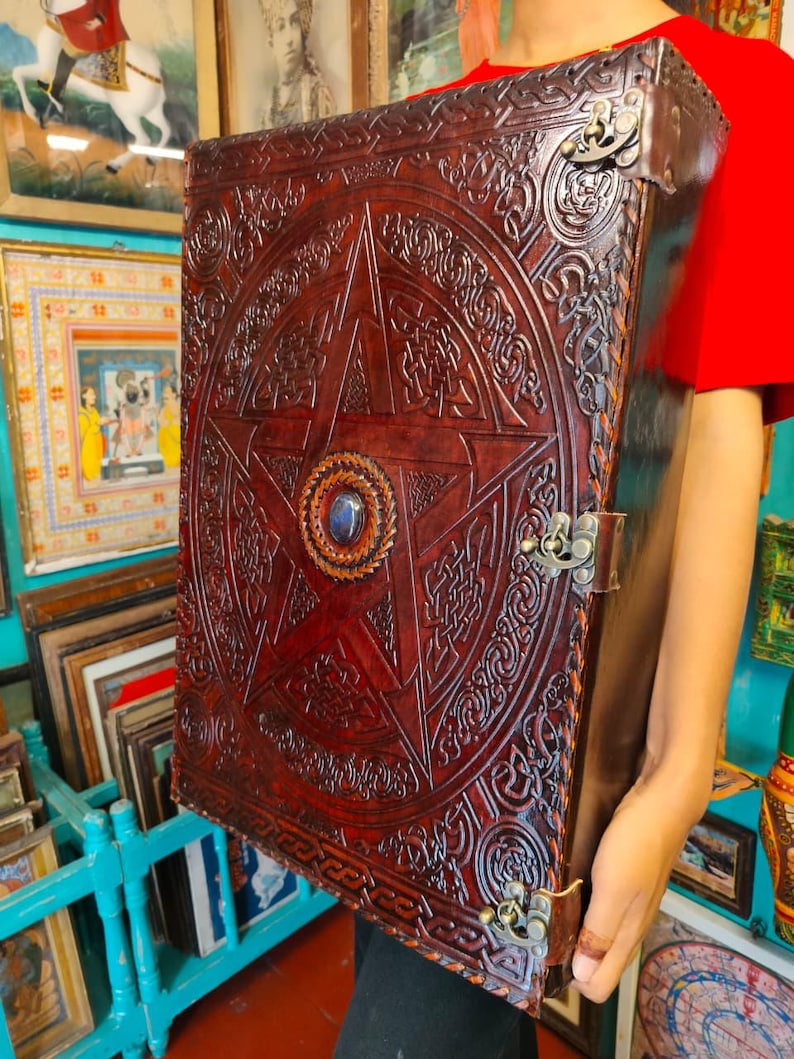 May include: Large, dark brown leather-bound book with a prominent embossed pentagram design. The cover features intricate Celtic knotwork and a central blue gemstone. Metal clasps and corner protectors add to the antique aesthetic.