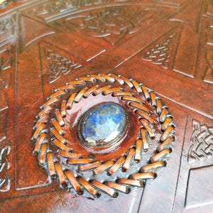 May include: Close-up of a brown leather item with intricate Celtic knotwork and a central, oval-shaped blue gemstone. The gemstone is surrounded by a stitched, circular border, adding a decorative element to the design.