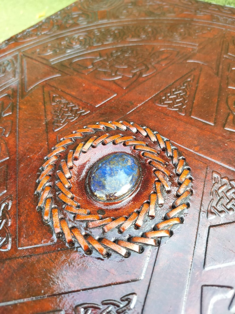 May include: Close-up of a brown leather item with intricate Celtic knotwork and a central, oval-shaped blue gemstone. The gemstone is surrounded by a stitched, circular border, adding a decorative element to the design.
