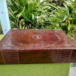 May include: A rectangular, dark brown leather box with intricate embossed designs. The box features a central starburst pattern and a circular jewel. The sides are stitched with a crisscross pattern. Decorative metal accents adorn the corners.