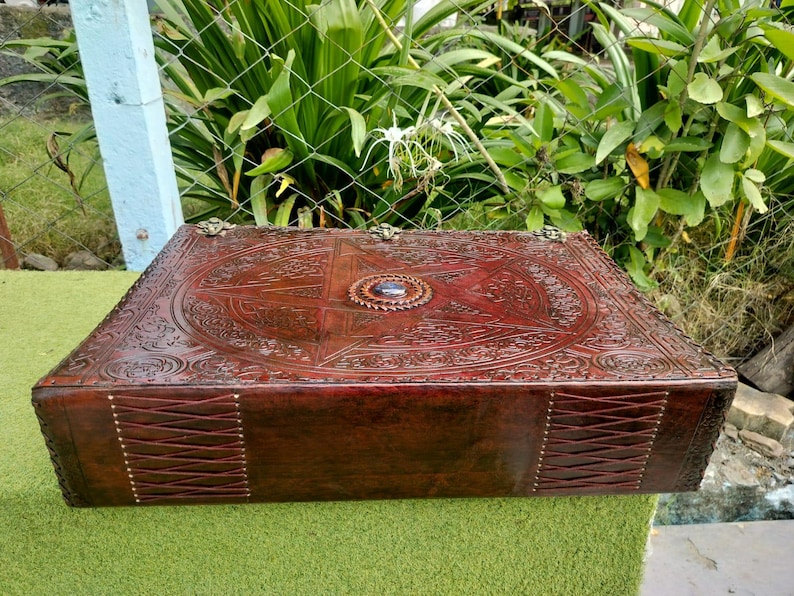 May include: A rectangular, dark brown leather box with intricate embossed designs. The box features a central starburst pattern and a circular jewel. The sides are stitched with a crisscross pattern. Decorative metal accents adorn the corners.