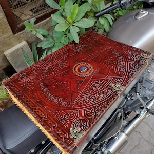 May include: A large, red leather-bound book with intricate embossed designs, including a central star and circular emblem. The book features brass corner accents and a blue gemstone. The book is resting on a motorcycle seat.