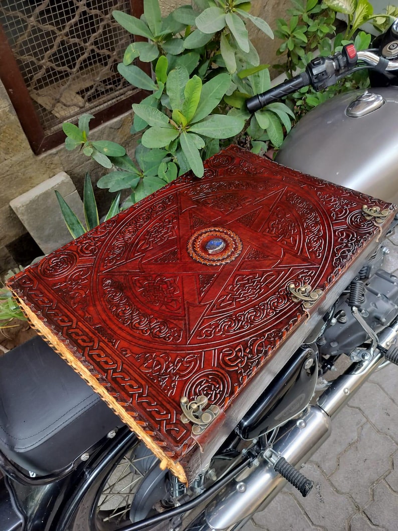 May include: A large, red leather-bound book with intricate embossed designs, including a central star and circular emblem. The book features brass corner accents and a blue gemstone. The book is resting on a motorcycle seat.