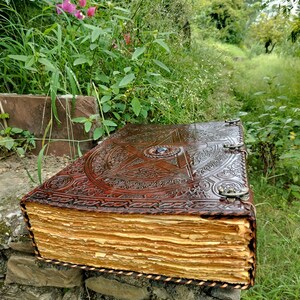 May include: A large, antique-style leather-bound journal with intricate embossed designs and a central star emblem. The aged pages have a golden hue, and the journal is secured with metal clasps. The journal is resting on a stone wall, surrounded by greenery.