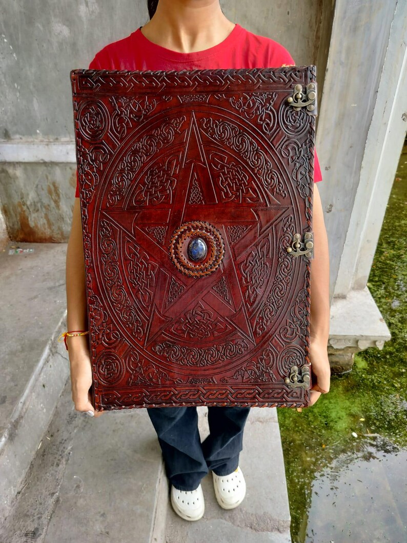 May include: A large, rectangular, burgundy leather-bound journal with an embossed pentagram design. The cover features intricate Celtic knotwork and a central, circular embellishment with a blue stone. Antique brass clasps secure the journal.