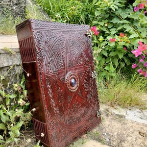 May include: A large, ornate, dark brown leather-bound book with intricate embossed designs. The cover features a central pentagram with a blue stone, surrounded by Celtic knotwork and floral patterns. Metal clasps secure the book.
