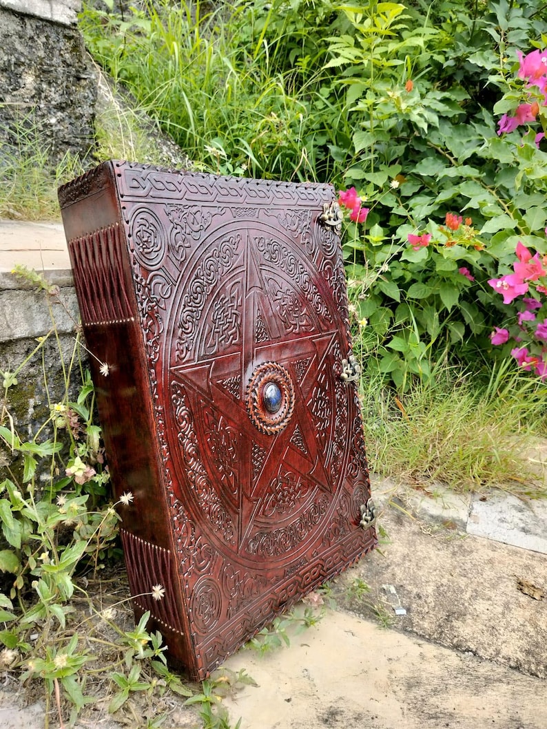 May include: A large, ornate, dark brown leather-bound book with intricate embossed designs. The cover features a central pentagram with a blue stone, surrounded by Celtic knotwork and floral patterns. Metal clasps secure the book.