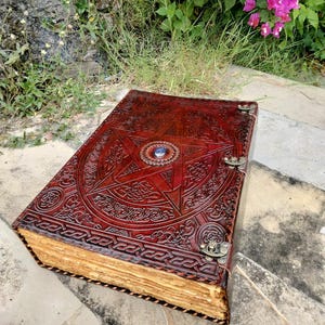 May include: A large, open, leather-bound journal with a detailed embossed design. The cover is a deep reddish-brown with a central pentagram and intricate patterns. The aged pages are visible, and the journal is secured with metal clasps.
