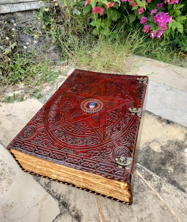 May include: A large, open, leather-bound journal with a detailed embossed design. The cover is a deep reddish-brown with a central pentagram and intricate patterns. The aged pages are visible, and the journal is secured with metal clasps.