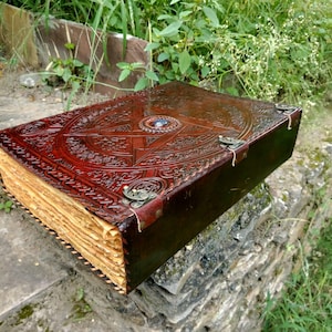May include: A large, antique-style leather-bound book with intricate embossed designs. The cover is a rich brown color, featuring a central starburst pattern and decorative metal clasps. The aged pages have a golden hue.