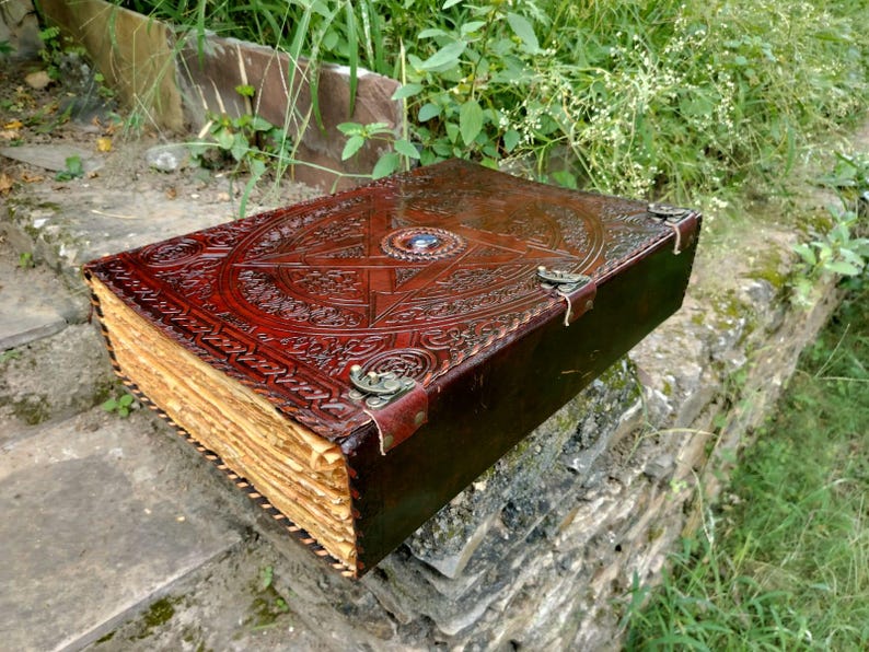 May include: A large, antique-style leather-bound book with intricate embossed designs. The cover is a rich brown color, featuring a central starburst pattern and decorative metal clasps. The aged pages have a golden hue.
