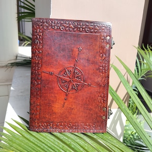 Puede incluir: Un diario de cuero marrón con un diseño de brújula en relieve en la portada. El diario tiene un borde decorativo y cierres metálicos.