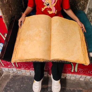 May include: An open, oversized book with aged, yellowed pages. The book's edges are rough and textured, suggesting a vintage or antique aesthetic. The book is held open, showcasing the blank pages.