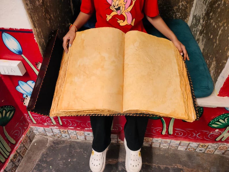 May include: An open, oversized book with aged, yellowed pages. The book's edges are rough and textured, suggesting a vintage or antique aesthetic. The book is held open, showcasing the blank pages.