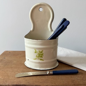 May include: A cream-coloured enamel wall-mounted container with a green floral motif on the front. The container features a rounded back panel with a hole for hanging. Inside are several dark blue handled utensils, with one matching knife.