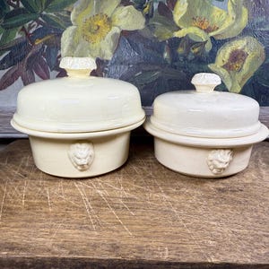 May include: Two cream-coloured ceramic lidded dishes. Each dish features a lion's head detail. The dishes are on a wooden surface, with a floral painting in the background.