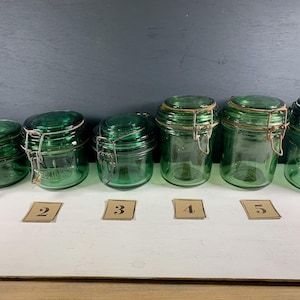 May include: Six green glass jars with metal lids and rubber seals. The jars are lined up in a row on a white surface. Each jar has a number written on a brown paper tag in front of it.
