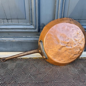 May include: A copper frying pan with a long handle. The pan has a round cooking surface and a dark metal handle. The pan is resting on a patterned surface in front of a blue door.